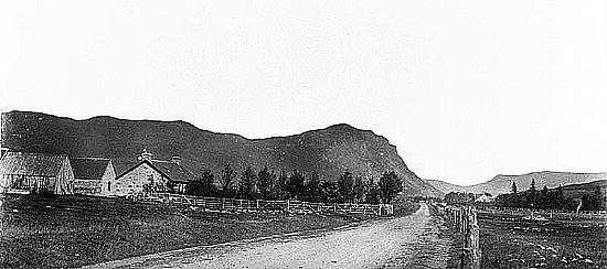 Black and white photo of a mountain with cottages, a fence and a dirt track in the foreground