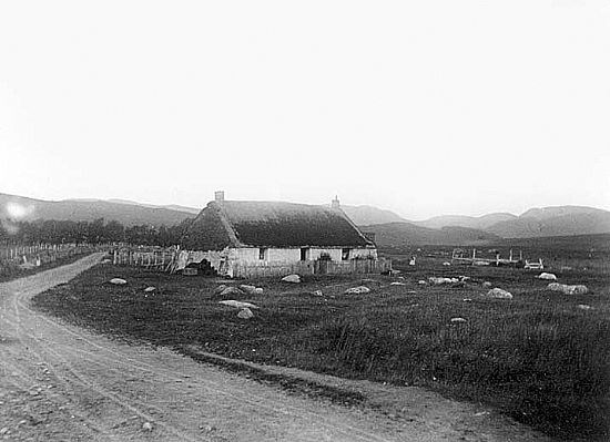  Black and white photo of thatched cottage beside a dirt track with mountains in the background
